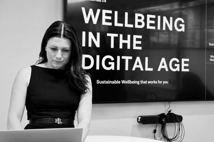 Woman working on a laptop in front of a screen displaying 'Wellbeing in the Digital Age'.