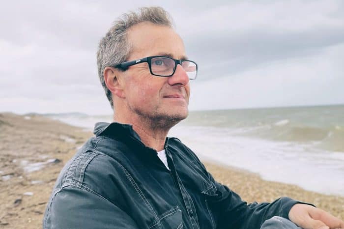 Man with glasses on a beach with waves and cloudy sky in the background.